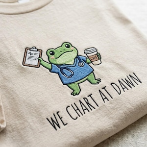 May include: Beige t-shirt featuring an embroidered cartoon frog wearing a blue medical scrub top and stethoscope, holding a clipboard and a coffee cup. The text "WE CHART AT DAWN" is embroidered below the frog.