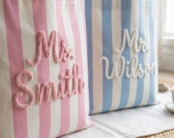 May include: Two striped tote bags, one pink and white, the other blue and white. The pink bag has "Ms. Smith" embroidered on it, and the blue bag has "Mr. Wilson" embroidered on it. Both bags have matching handles.