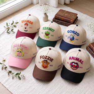 Puede incluir: Seis gorras de béisbol beige con personajes de los Muppets bordados. Cada gorra muestra un nombre, un año de creación y una visera de color. Los personajes incluyen a Beaker, Kermit, Gonzo, Miss Piggy, Fozzie y Animal, con viseras en burdeos, verde, azul, rosa, marrón y negro.