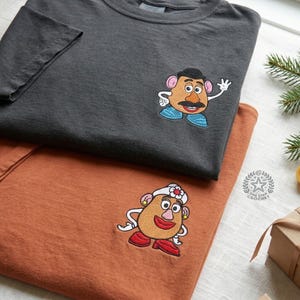 Pode incluir: Uma t-shirt cinzento-carvão com um bordado da personagem Mr. Potato Head e uma t-shirt cor de ferrugem com um bordado da personagem Mrs. Potato Head. Ambas as t-shirts têm mangas curtas e gola redonda com detalhes bordados precisos.