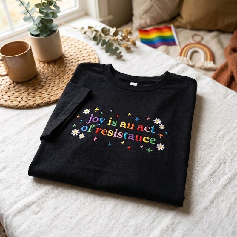 May include: Black t-shirt with the embroidered text "joy is an act of resistance" in colorful letters and floral accents. The shirt is folded on a white surface, with a mug and plant visible in the background.
