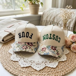 May include: Two beige trucker caps with floral patterned peaks. One cap has the word "FOUR" in black, the other has "CUSTOM" in green. The caps are displayed on a woven mat.