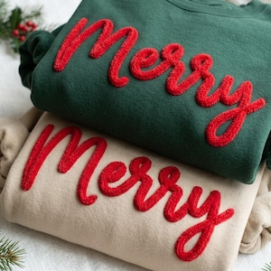 Op de afbeelding: Een donkergroen sweatshirt en een beige sweatshirt, beide voorzien van het woord Merry in rode, verhoogde, getextureerde letters in chenille-stijl op de voorzijde. De kledingstukken zijn opgevouwen en op elkaar gestapeld.
