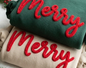 May include: A dark green sweatshirt and a beige sweatshirt, each featuring the word Merry in red, raised, textured chenille-style lettering across the front. The garments are folded and stacked, showcasing the soft fabric and bold script.