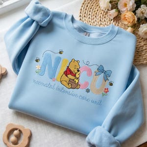 May include: A light blue sweatshirt with embroidered text and graphics. The word "NICU" is spelled out in colorful letters, with a Winnie the Pooh graphic. The phrase "neonatal intensive care unit" is embroidered below.