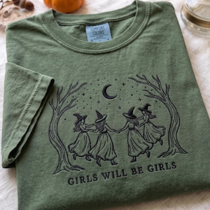 Peut inclure: Un t-shirt vert olive avec une broderie noire repr&eacute;sentant quatre silhouettes portant des chapeaux de sorci&egrave;re et des robes, dansant en cercle entre deux arbres sous un croissant de lune et des &eacute;toiles. Le texte GIRLS WILL BE GIRLS est brod&eacute;.