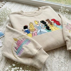May include: Beige sweatshirt featuring an embroidered design of several Disney princesses. The sleeve has an embroidered castle design. The princesses are depicted in their iconic dresses. The sweatshirt is made of a soft material.