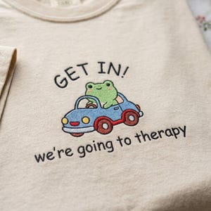 May include: Beige t-shirt with an embroidered design. The design features a green frog driving a blue car with red wheels. Text above the car reads "GET IN!" and below it, "we're going to therapy."