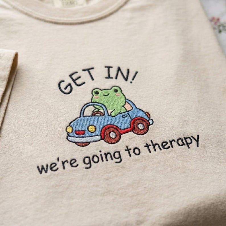 May include: Beige t-shirt with an embroidered design. The design features a green frog driving a blue car with red wheels. Text above the car reads "GET IN!" and below it, "we're going to therapy."