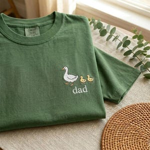 Puede incluir: Camiseta verde Comfort Colors con un dise&ntilde;o bordado de un pato blanco, dos patitos amarillos y la palabra "dad" en blanco. La camiseta est&aacute; doblada sobre una superficie neutra.