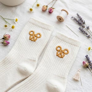 May include: A pair of cream-coloured ribbed socks featuring embroidered pretzel designs. The socks are presented on a white surface with dried flowers, chamomile, and sewing supplies. The pretzels are golden brown.