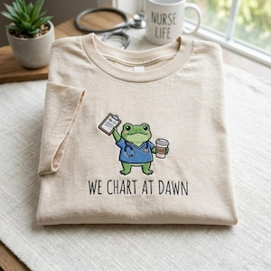 May include: Beige t-shirt featuring a cartoon frog in blue scrubs, holding a clipboard and a coffee cup. The text "WE CHART AT DAWN" is printed below the frog. A mug with "NURSE LIFE" is in the background.
