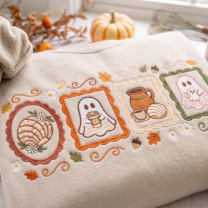 May include: A beige sweatshirt featuring detailed embroidery of four framed scenes. The designs include a concha bread, a ghost holding a coffee cup, a pitcher with a cup and bread, and a ghost holding a smaller ghost, all accented with leaves and flowers.