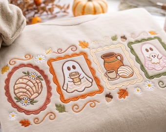 May include: A beige sweatshirt featuring detailed embroidery of four framed scenes. The designs include a concha bread, a ghost holding a coffee cup, a pitcher with a cup and bread, and a ghost holding a smaller ghost, all accented with leaves and flowers.