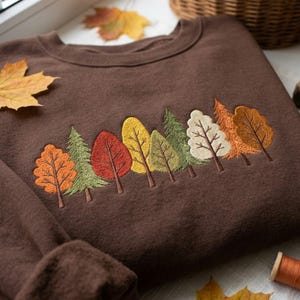 May include: A dark brown crewneck sweatshirt featuring an embroidered row of various trees in autumn colors including orange, green, red, yellow, and cream. The design depicts a forest scene with different leaf shapes and textures across the chest.