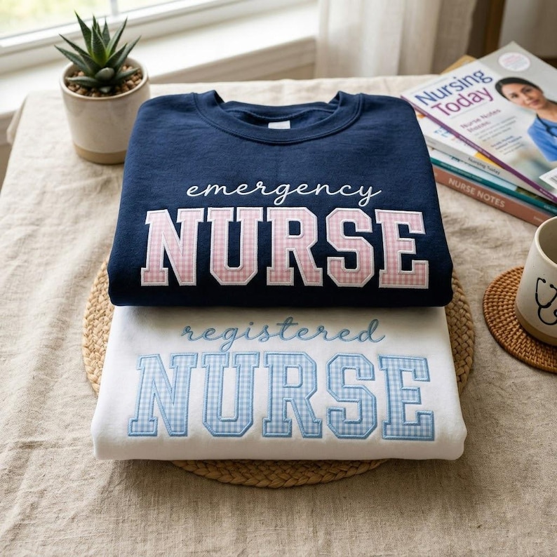 May include: Two sweatshirts, one navy blue and one white, are stacked on a woven mat. The navy sweatshirt says "emergency NURSE" in white script and pink gingham letters. The white sweatshirt says "registered NURSE" in blue script and gingham letters.