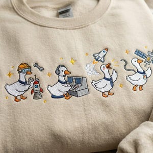 May include: Beige sweatshirt with an embroidered design of cartoon geese in space-themed roles. One goose wears a hard hat and works on a rocket, another operates a control panel, and others are near a rocket launch and satellite.