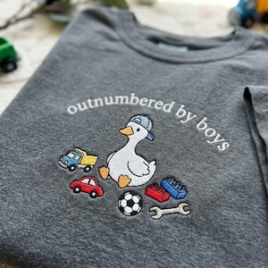 K&ouml;nnte beinhalten: Graues T-Shirt mit gesticktem Design. Es zeigt eine Ente mit blauer Kappe, umgeben von Spielzeugautos, Bausteinen, einem Fu&szlig;ball und einem Schraubenschl&uuml;ssel. Der Text "outnumbered by boys" ist &uuml;ber der Ente angeordnet.