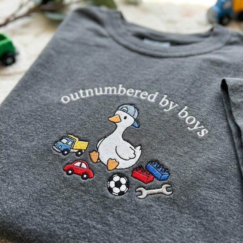 K&ouml;nnte beinhalten: Graues T-Shirt mit gesticktem Design. Es zeigt eine Ente mit blauer Kappe, umgeben von Spielzeugautos, Bausteinen, einem Fu&szlig;ball und einem Schraubenschl&uuml;ssel. Der Text "outnumbered by boys" ist &uuml;ber der Ente angeordnet.