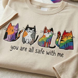 May include: A beige t-shirt featuring an embroidered design of four cats with distinct colourful patterns, including a rainbow cat. Below the cats, the text "you are all safe with me" is embroidered in black thread across the chest area.