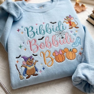 May include: A light blue sweatshirt featuring embroidered text that reads Bibbidi Bobbidi Boo. The design includes two cartoon mice, a spider web, bats, stars, a glass slipper, and two pumpkins, one with a pink bow, all in colorful thread.