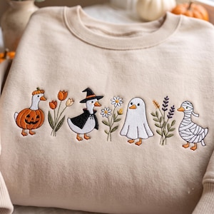 May include: A beige crewneck sweatshirt featuring an embroidered horizontal row of three ducks in costumes, a ghost, and floral stems. The designs include a duck in a pumpkin suit, a witch duck, a ghost, and a mummy duck, interspersed with flowers.