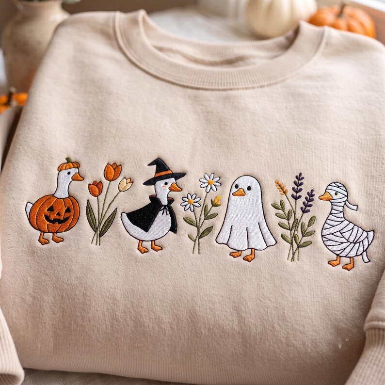 May include: A beige crewneck sweatshirt featuring an embroidered horizontal row of three ducks in costumes, a ghost, and floral stems. The designs include a duck in a pumpkin suit, a witch duck, a ghost, and a mummy duck, interspersed with flowers.