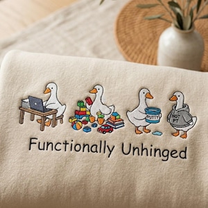 Könnte beinhalten: Beiges Sweatshirt mit einem gestickten Design, das vier Cartoon-Gänse zeigt. Eine Gans sitzt an einem Laptop, eine andere mit Spielzeug, eine mit Kitt und die letzte in einem Kapuzenpullover. Der Text "Functionally Unhinged" steht darunter.