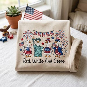 May include: A folded beige sweatshirt featuring an embroidered design of patriotic geese dressed in red, white, and blue attire, including a Statue of Liberty goose. The text "Red, White And Goose" is printed below the design.