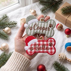 May include: Two bone-shaped pet ornaments with metal rings. The top ornament is sage green and white striped with a green Santa hat and the name Cooper. The bottom ornament is red and white checkered with a red Santa hat and the name Bella.