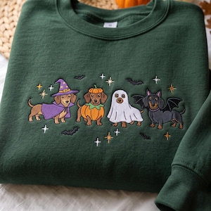 May include: A dark forest green crewneck sweatshirt featuring an embroidered design of four dachshunds in Halloween costumes. The dogs are dressed as a wizard, a pumpkin, a ghost, and a bat, surrounded by small embroidered stars and black bats.