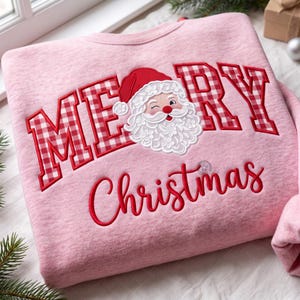 May include: A light pink sweatshirt featuring the text MERY in red and white gingham block letters, with a white embroidered Santa Claus face replacing the letter A. Below, the word Christmas is embroidered in red cursive script.