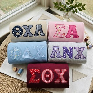 May include: Five folded sweatshirts in beige, pink, light blue, gray, and maroon, each featuring large Greek letter appliqués with varied patterns including stars, gingham, polka dots, and floral prints, arranged on a textured surface.