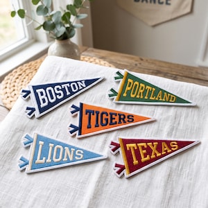 May include: Five triangular felt pennants featuring embroidered text. The pennants are navy blue with white text reading BOSTON, green with yellow text reading PORTLAND, orange with navy text reading TIGERS, light blue with white text reading LIONS, and maroon with yellow text reading TEXAS.