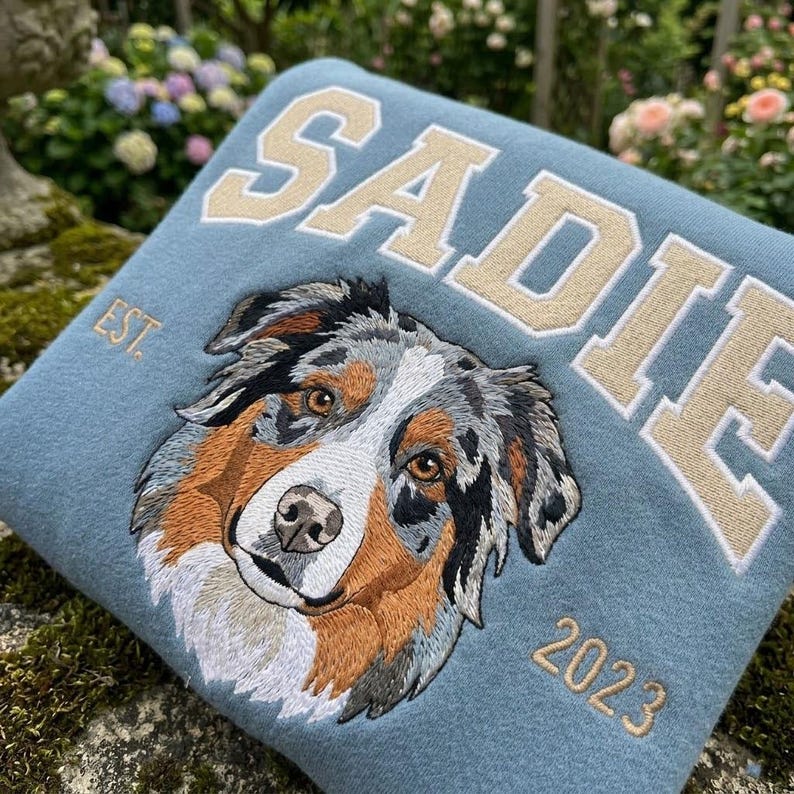 May include: A light blue sweatshirt with the name "SADIE" embroidered in a cream-colored collegiate font. Below the name is an embroidered portrait of a dog with brown, gray, and white fur. The year "2023" is embroidered below the dog.