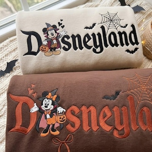 May include: Two folded sweatshirts, one beige and one brown, featuring embroidered Disneyland text, Mickey Mouse in a witch hat holding a pumpkin, a spider web, and bats. The beige sweatshirt has black embroidery, while the brown sweatshirt features orange and black embroidery.
