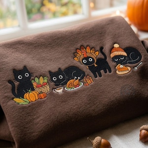 Puede incluir: Una sudadera marrón con un diseño bordado de cuatro gatos negros. Los gatos aparecen con elementos de temática otoñal, como una cesta de verduras, un pavo asado en una fuente, un disfraz de pavo y un trozo de tarta de calabaza.