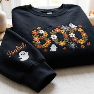 May include: A black crewneck sweatshirt featuring embroidered floral letters spelling BOO in orange, yellow, and white. The design includes small white ghosts, spiderwebs, and leaves. The sleeve is embroidered with the name Isabel and a ghost.