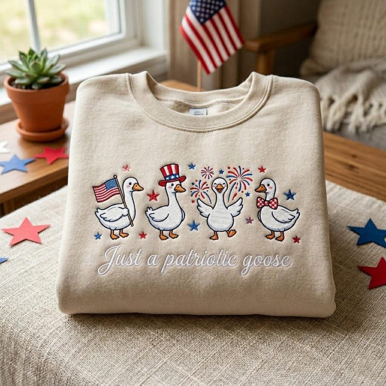 May include: A beige sweatshirt with an embroidered design of four cartoon geese celebrating Independence Day. One goose holds an American flag, another wears a patriotic hat, and the others are surrounded by fireworks. The text "Just a patriotic goose" is embroidered below.
