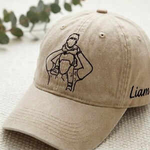 May include: A tan baseball cap with a black embroidered design of a person carrying a child on their shoulders. The name "Liam" is printed on the side. The cap has a curved brim and a button on top.