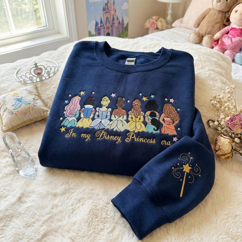 May include: Navy blue sweatshirt with embroidered Disney princess design. Features a row of princess figures with various hair colors and gowns. The text "In my Disney Princess era" is embroidered below the princesses. A gold embroidered wand is on the sleeve.