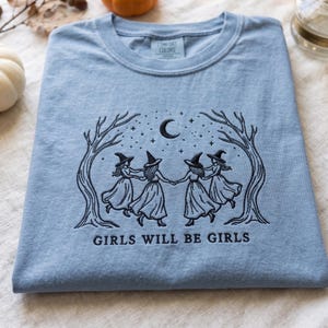 Peut inclure: Un t-shirt bleu clair avec une broderie noire repr&eacute;sentant quatre silhouettes portant des chapeaux de sorci&egrave;re, se tenant par la main sous un croissant de lune et des &eacute;toiles, encadr&eacute;es par deux arbres nus. Le texte GIRLS WILL BE GIRLS est brod&eacute;.