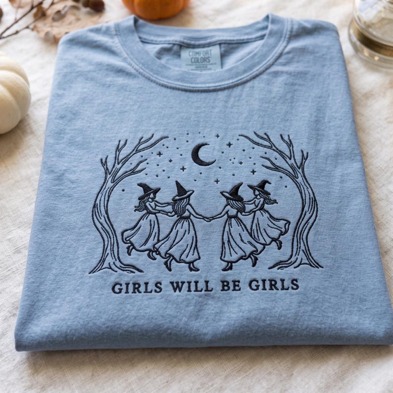 Peut inclure: Un t-shirt bleu clair avec une broderie noire repr&eacute;sentant quatre silhouettes portant des chapeaux de sorci&egrave;re, se tenant par la main sous un croissant de lune et des &eacute;toiles, encadr&eacute;es par deux arbres nus. Le texte GIRLS WILL BE GIRLS est brod&eacute;.