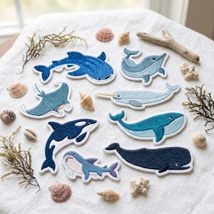 May include: A collection of embroidered fabric patches featuring various marine animals including a whale shark, dolphin, manta ray, narwhal, blue whale, orca, and sperm whale, arranged on a white fabric surface with scattered seashells and driftwood.