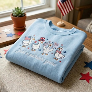 May include: Light blue sweatshirt with embroidered patriotic geese, each adorned with festive accessories like an American flag, a top hat, and a bow tie. The text "Just a patriotic goose" is embroidered below the geese.
