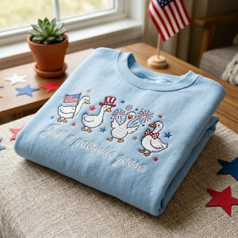 May include: Light blue sweatshirt with embroidered patriotic geese, each adorned with festive accessories like an American flag, a top hat, and a bow tie. The text "Just a patriotic goose" is embroidered below the geese.