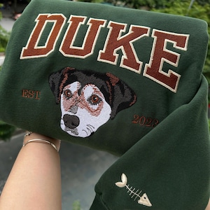 May include: Dark green sweatshirt with the name "DUKE" embroidered in brown block letters. Below the name is an embroidered dog portrait with the text "EST 2023". The sleeve features a fishbone design.