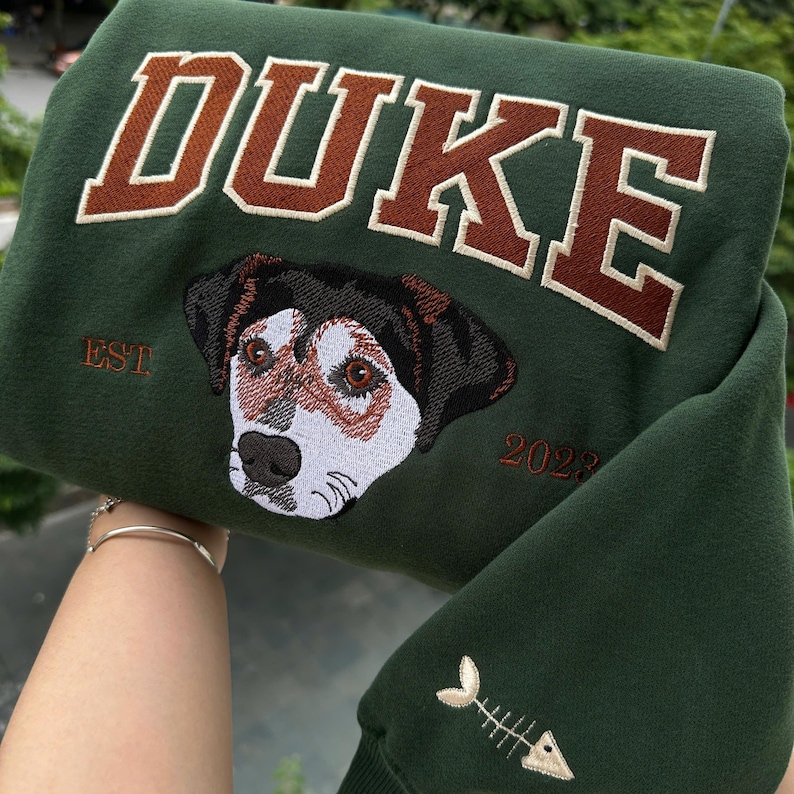 May include: Dark green sweatshirt with the name "DUKE" embroidered in brown block letters. Below the name is an embroidered dog portrait with the text "EST 2023". The sleeve features a fishbone design.