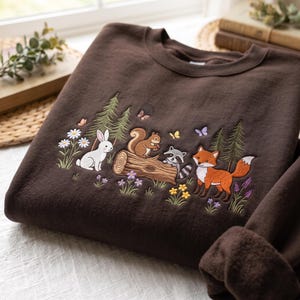 May include: A dark brown crewneck sweatshirt featuring an embroidered forest scene. The design includes a white rabbit, a squirrel on a log, a raccoon, and an orange fox, surrounded by pine trees, colorful wildflowers, and small butterflies.