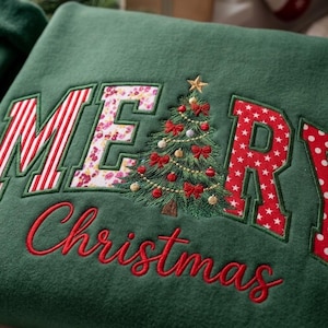 Op de afbeelding: Een donkergroene sweater met de geborduurde tekst MERRY, waarbij een kerstboom de letter E vervangt. De letters M, R en Y hebben rood-witte patronen, terwijl de E een bloemenprint heeft. Het woord Christmas is eronder geborduurd.